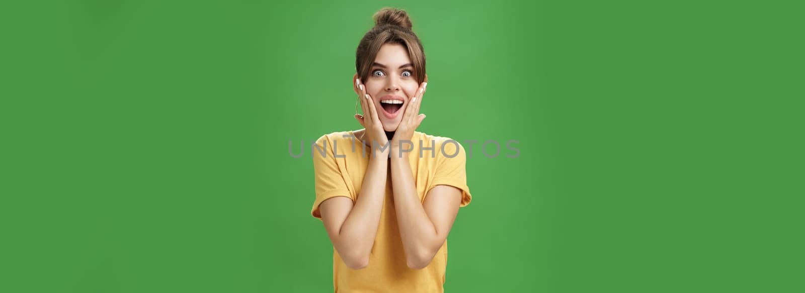 Indoor shot of excited and surprised energized attractive glamorous female in yellow t-shirt touching cheeks from amazement smiling broadly astonished posing over green background by Benzoix