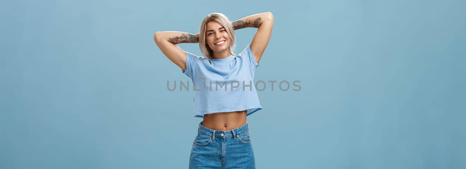 Thanks god it weekends. Relaxed lazy and attractive young stylish woman with fari hair tattoos, pierced belly stretching with delight holding hands behind head in carefree pose, smiling over blue wall by Benzoix