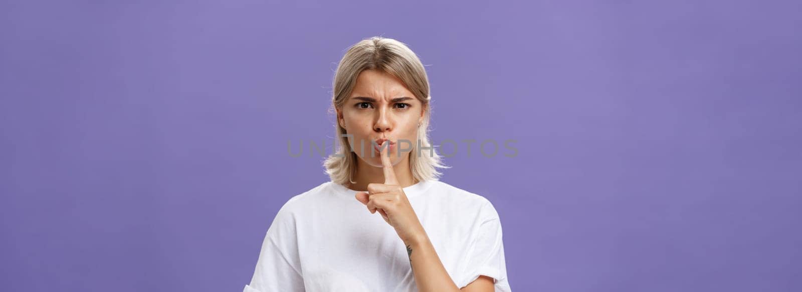 Close-up shot of serious strict good-looking caucasian female with blond hair shushing at camera displeased and disappointed frowning holding index finger over folded lips in shush sign by Benzoix
