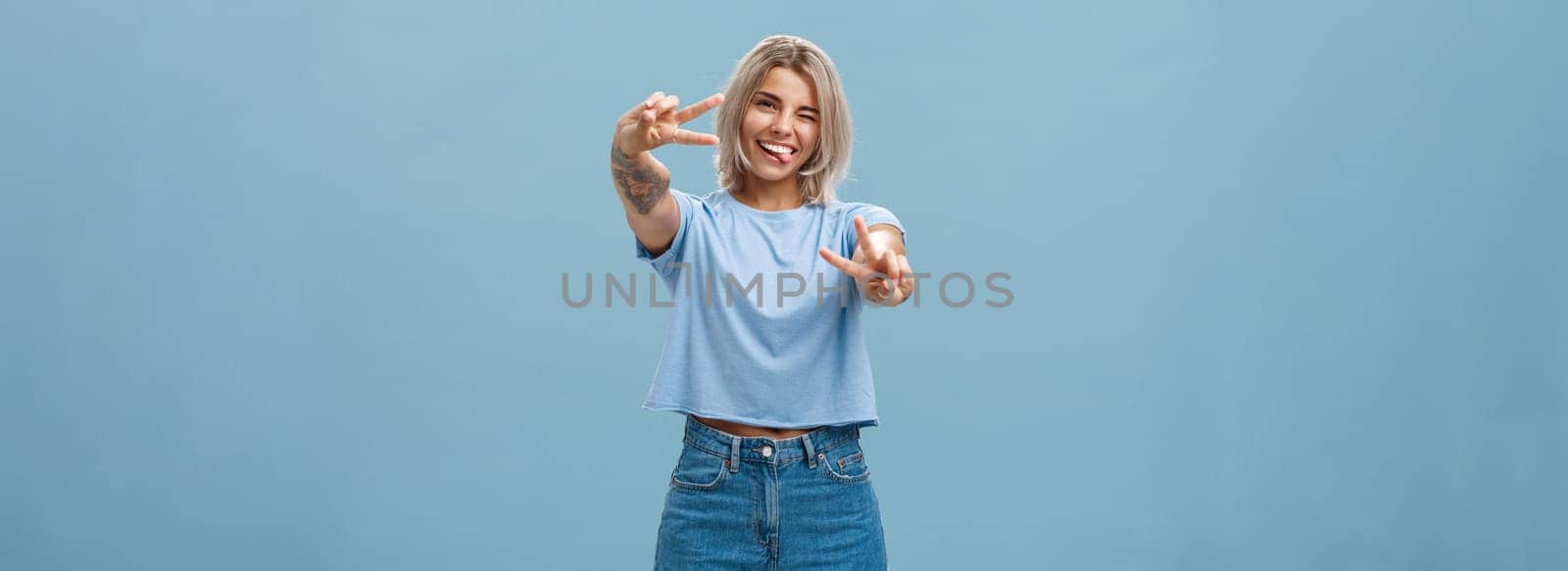 Beautiful tattooed girl enjoying weekends sticking out tongue joyfully winking at camera and smiling showing peace or victory gesture with pulled arms feeling happy posing over blue background. Copy space