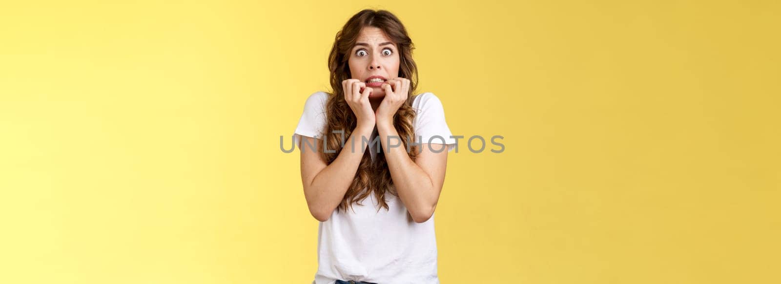 Scared timid insecure unconfident european girl curly haircut hold hands face biting fingers look frightened shocked standing stupor speechless shaking fear terrified yellow background.