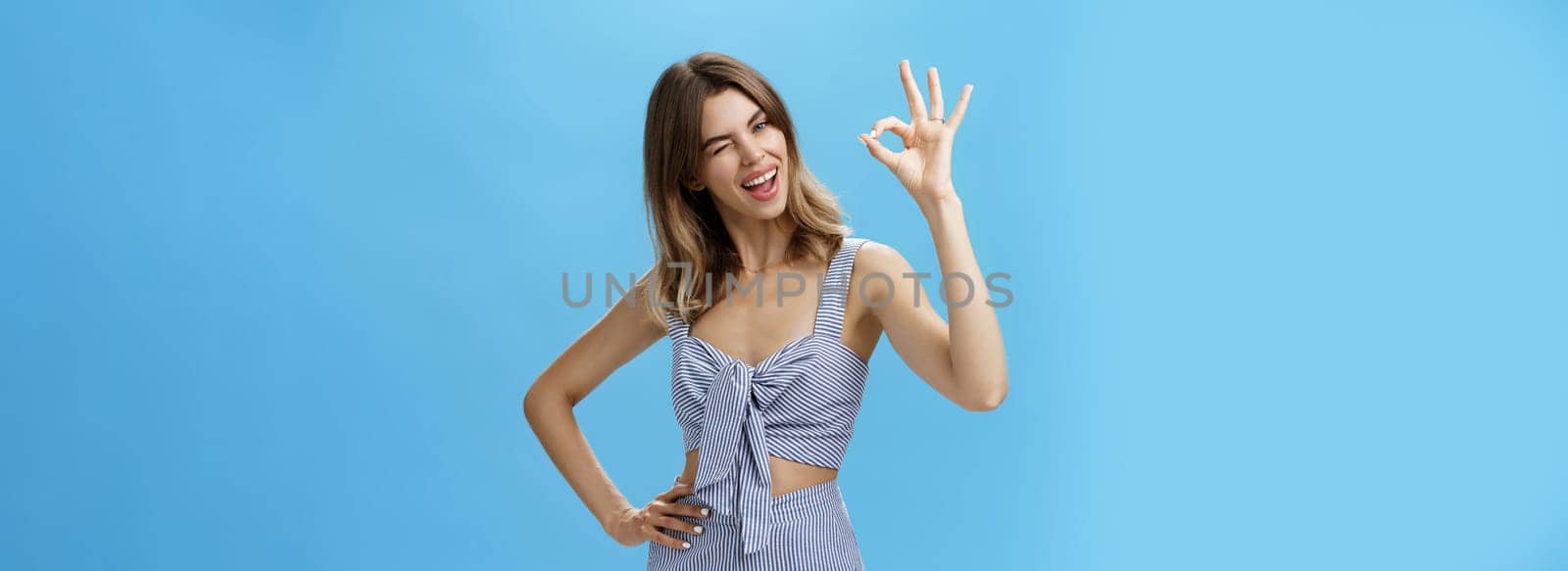 Cute charismatic woman with gap teeth showing okay gesture tilting head joyfully, holding hand on waist and winking with self-assured expression reassuring friend everything goes on plan. Body language concept