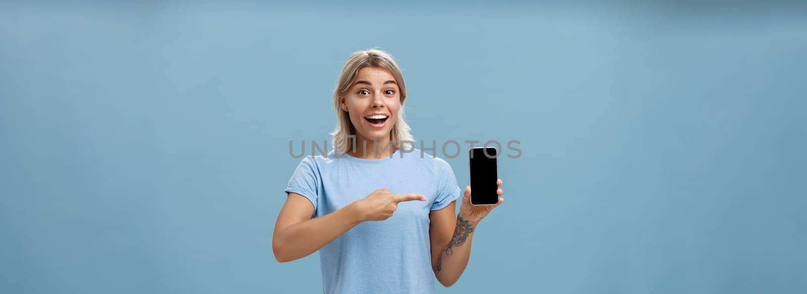 Lifestyle. Waist-up shot of thrilled and impressed good-looking female student in casual t-shirt smiling joyfully pointing at smartphone screen showing awesome place via internet to friend over blue background.