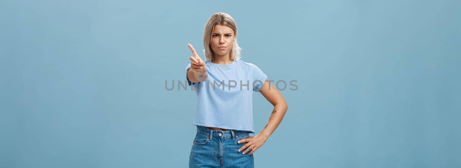 You better not do it. Serious-looking displeased strict elder sister with fair hair and tanned skin frowning holding hand on waist shaking index finger in forbid or stop gesture over blue background.