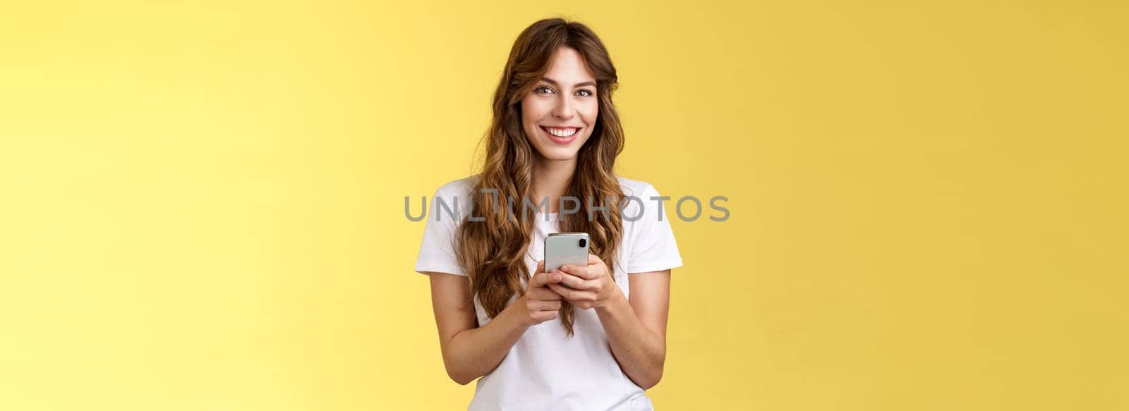 Enthusiastic charming sociable young girl messaging friend sending photos social media hold smartphone look camera happily friendly smiling stand yellow background casual outfit. Technology concept
