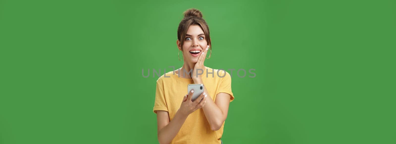 Surprised touched and impressed beautiful european girl with diastema smiling broadly touching cheek from amazement and joy holding smartphone looking happy at camera after reading cool news. Technology concept