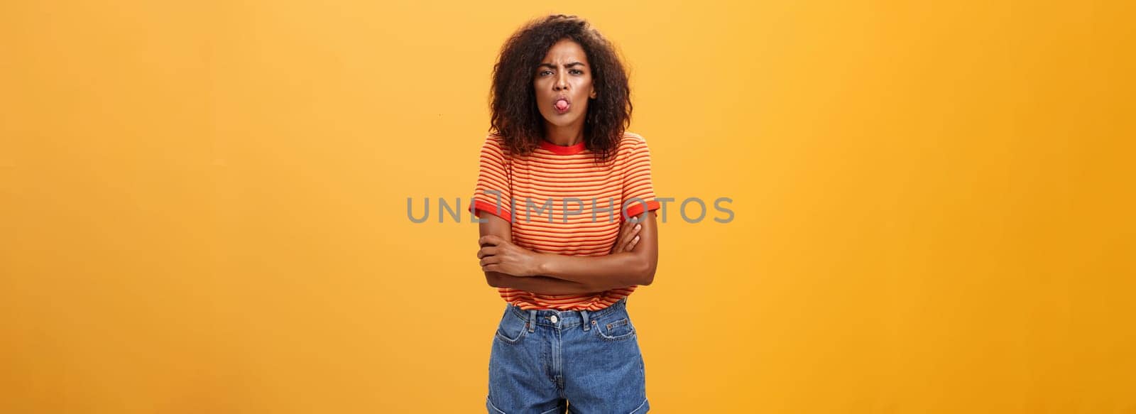 Immature girl showing bad side of character. Portrait of childish offended or displeased young African-American woman with curly hair showing tongue crossing arms on chest over orange background.