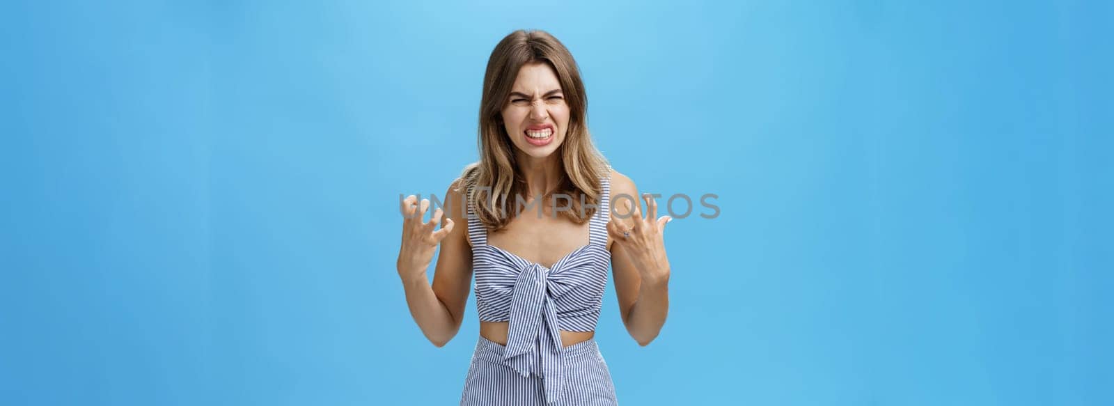 Indoor shot of irritated moody woman having mental breakdown feeling annoyed and pressured squeezing hands in fists from anger wrinkling nose in hate standing hateful over blue background by Benzoix