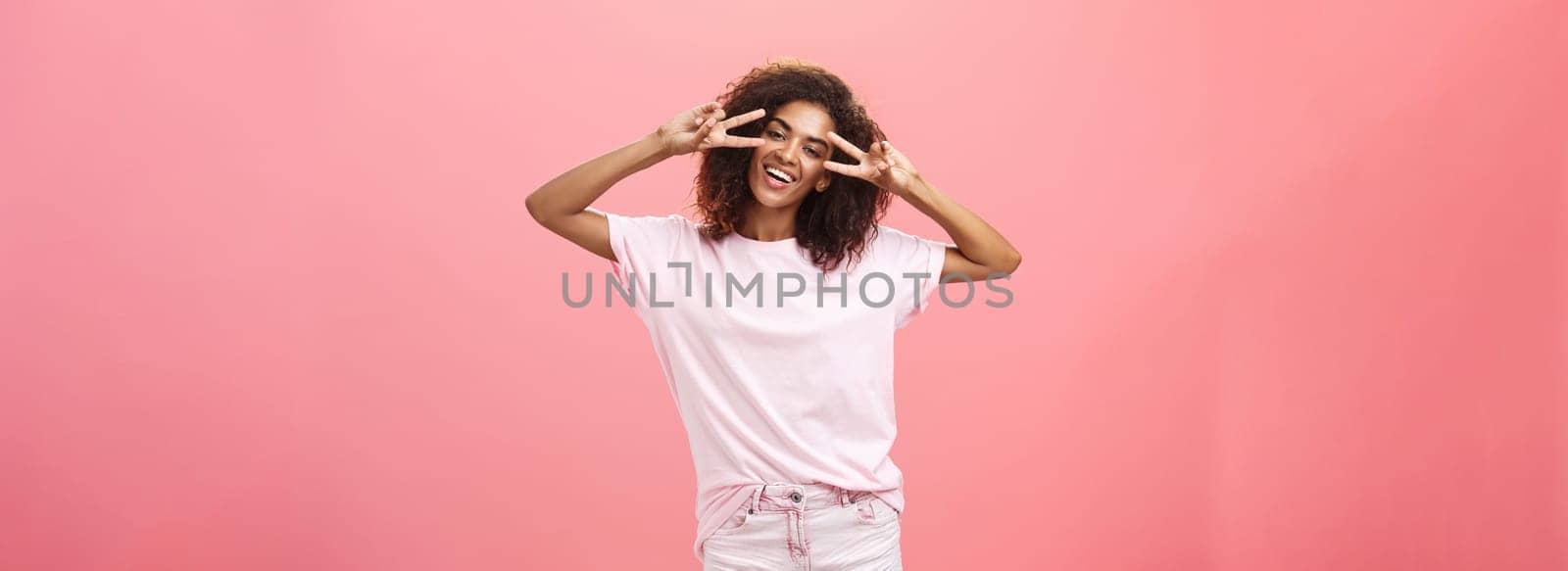 Indoor shot of charismatic playful dark-skinned woman with afro hairstyle tilting head showing peace or victory signs over eyes and smiling feeling amused chilling on awesome party over pink wall by Benzoix