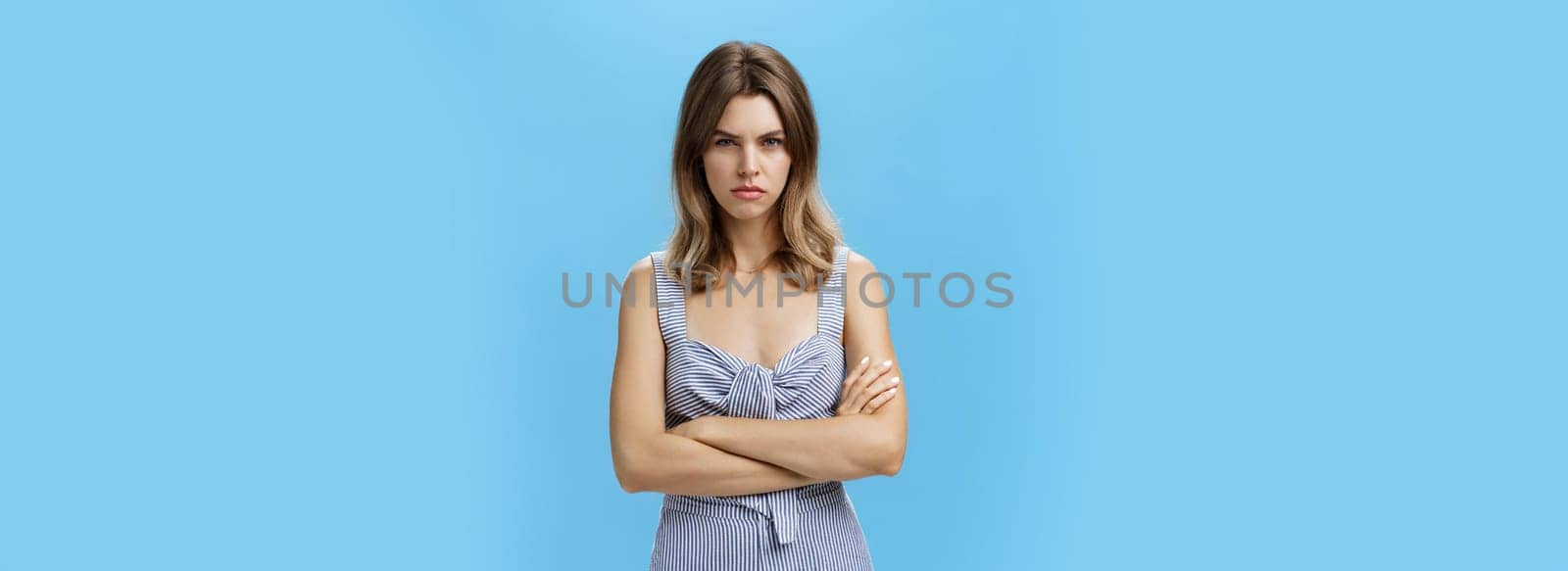 Woman waiting time for revenge. Portrait of intense offended and moody confident adult woman in cute dress frowing and sulking, holding hands crossed on chest from insult and jealousy over blue wall by Benzoix