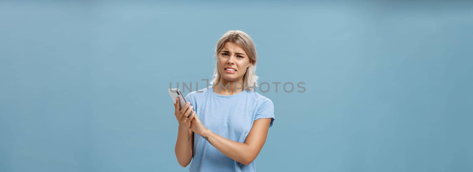 Woman cannot stand when mom shouts during phone call. Intense displeased young european blonde with tanned skin clenching teeth and frowning covering microphone of smartphone over blue wall by Benzoix