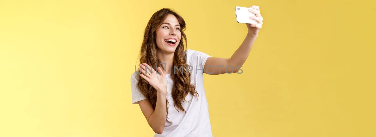 Friendly lively good-looking cheerful feminine girl extend arm hold smartphone record video blog waving front camera smiling broadly talking fans videocalling taking selfie yellow background. Lifestyle.