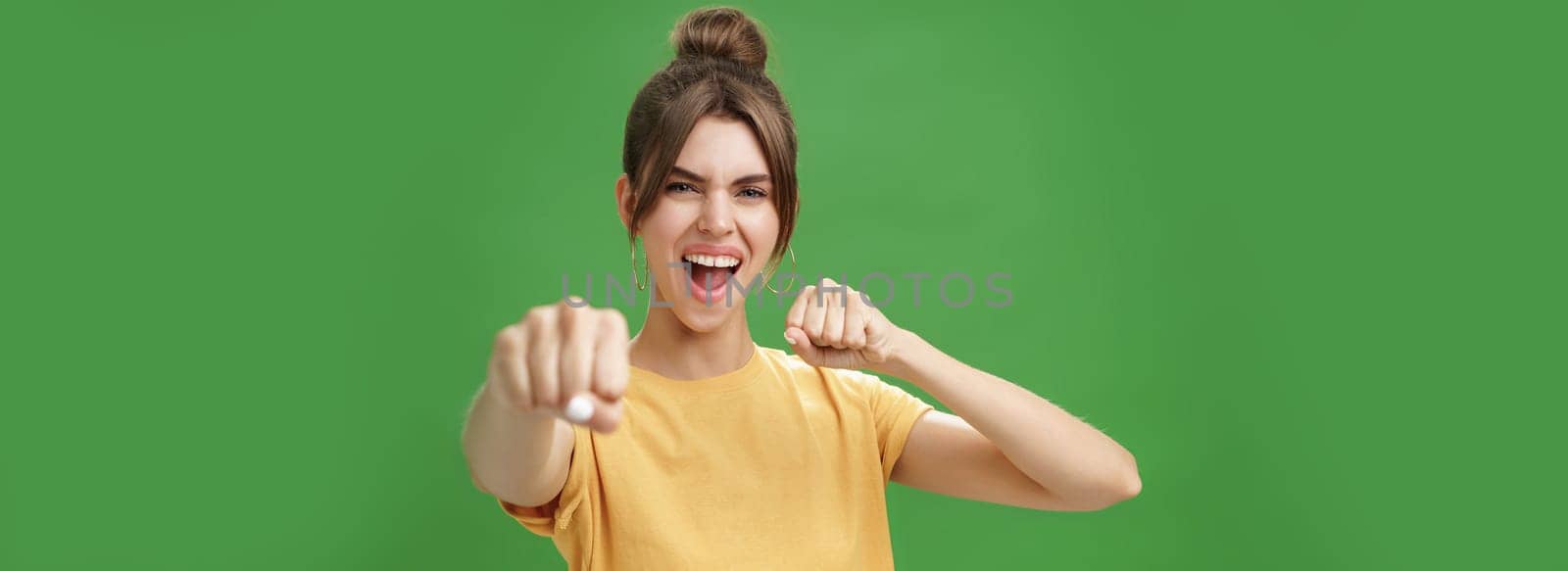 Cute female rebel in yellow t-shirt with gap teeth pulling fist towards camera as if showing fighting skills yelling daring and excited standing over green background smiling acting like boxer by Benzoix
