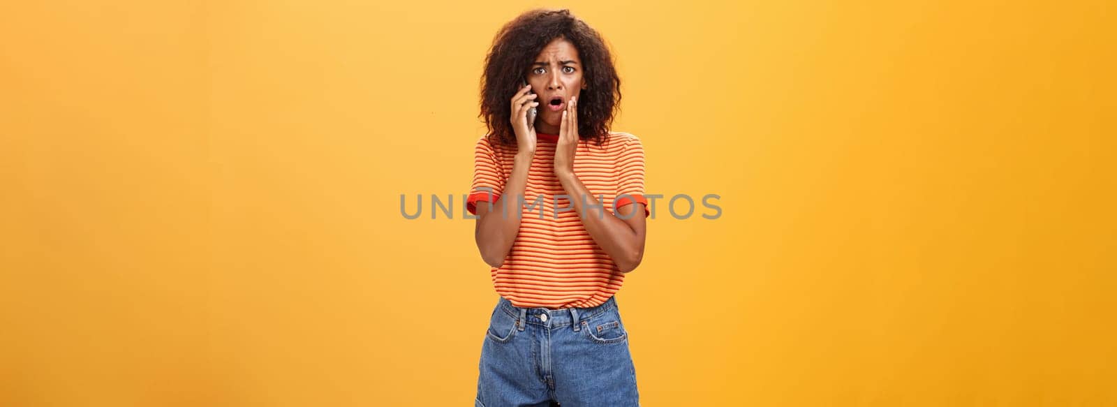 Woman receiving disturbing call feeling emapthy and sorry for poor friend getting in trouble holding cellphone near ear touching cheek concerned frowning standing sad, worried over orange background by Benzoix