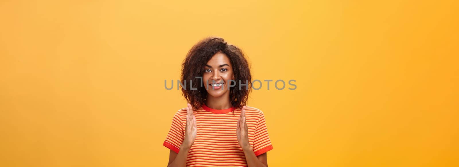 Portrait of charming friendly-looking african american woman with afro hairstyle explaining story with hands and gestures shaping object, holding palms vertical over chest and smiling at camera by Benzoix