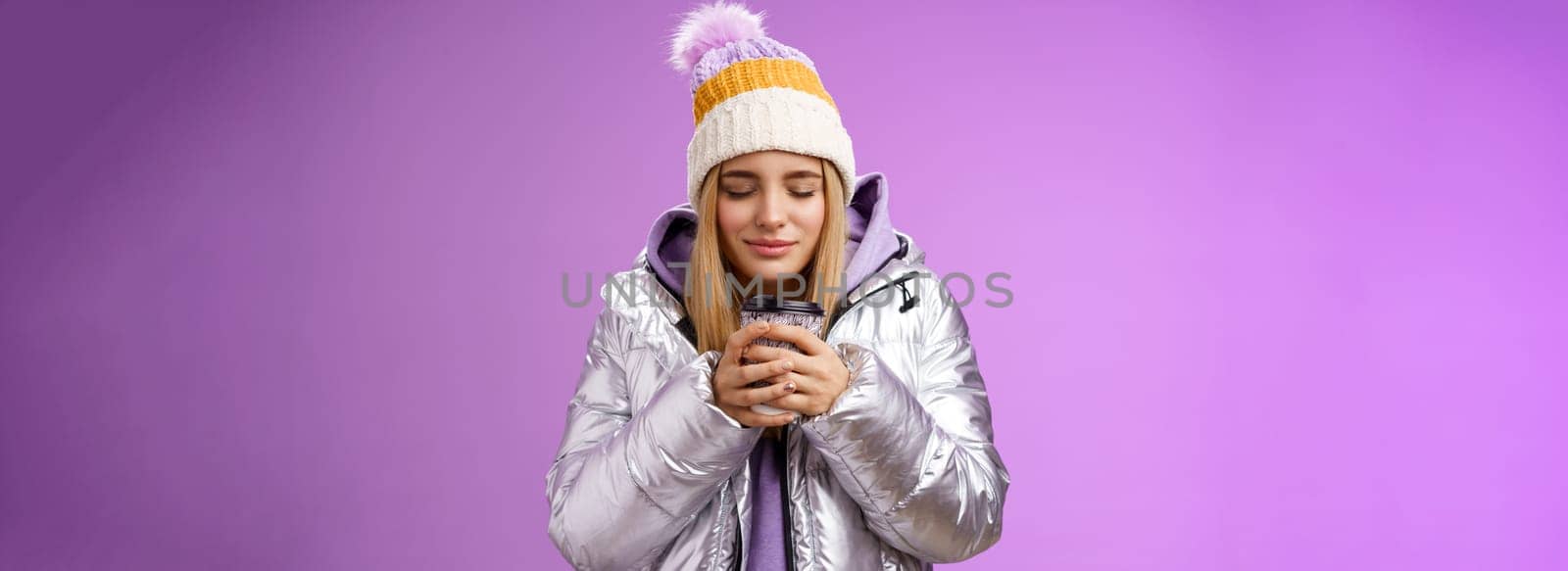 Charming tender romantic blond girl sniffing nice smell warm tasty coffee holding take-away cup warm hands close eyes look delighted wearing winter jacket hat enjoy vacation, purple background by Benzoix