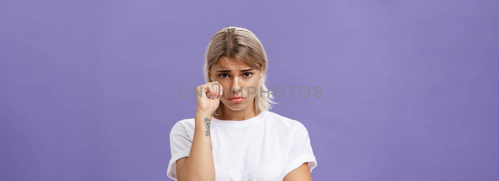 Studio shot of offended sad and timid silly woman with blond hairstyle frowning looking from under forehead holding fist near eye as if whiping teardrop being upset over purple background. Emotions concept