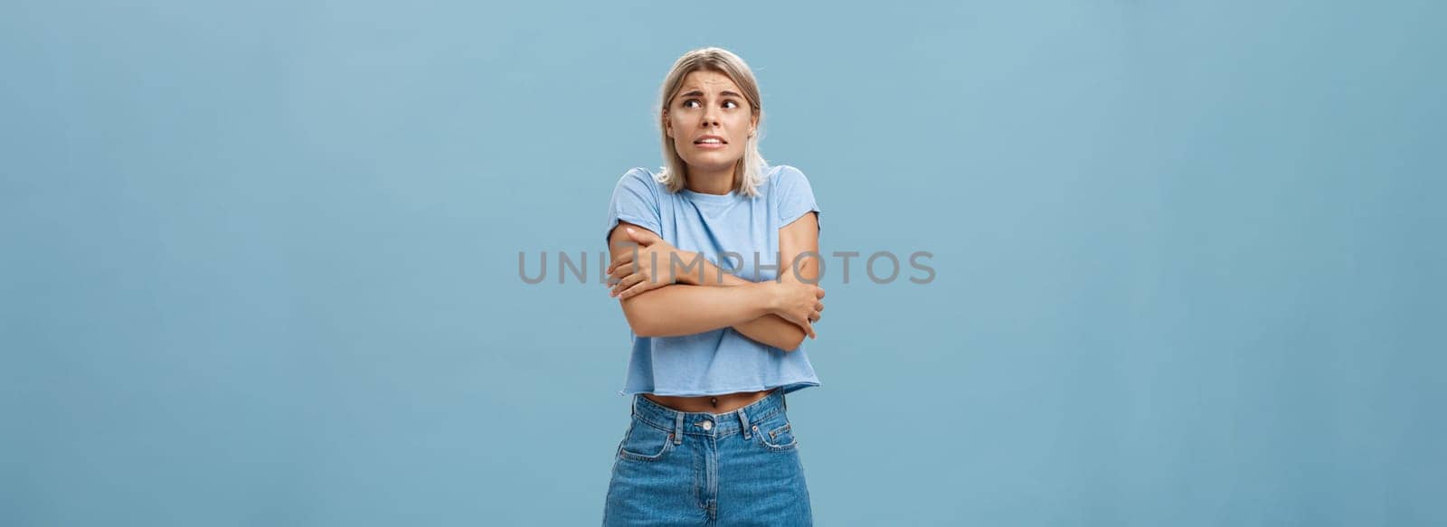 Turn off air-cooler I am freezing in summer. Displeased cute caucasian blonde girl in trendy t-shirt hugging herself while trembling from cold shaking gazing right with worried look over blue wall.