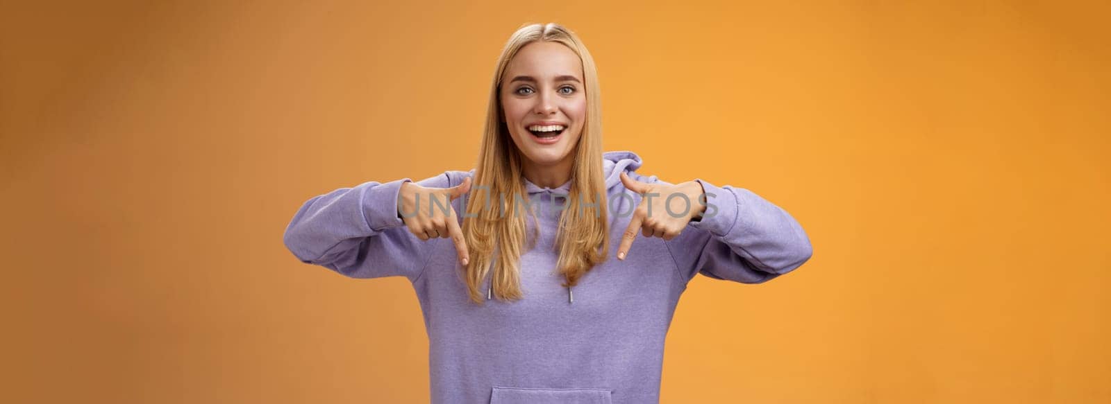 Amused joyful nice blond girlfriend pointing down present cool new product smiling broadly recommending try check out standing orange background happily grinning in hoodie. Advertisement concept