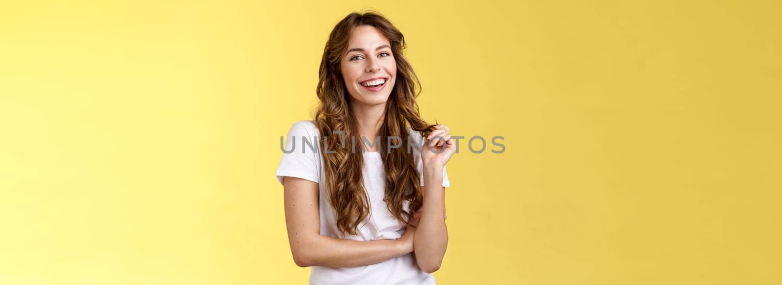 Lively joyful happy cute brunette curly long hairstyle playing curl laughing joyfully having funny amusing conversation smiling toothy white perfect smile stand yellow background relaxed.