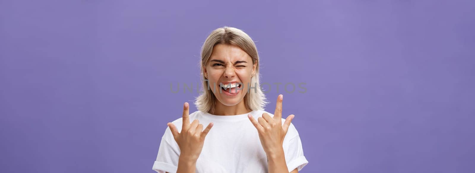 Gonna rock. Portrait of joyful happy good-looking stylish woman with blond medium haircut winking smiling and sticking out tongue while showing rock-n-roll gesture with both hands over purple wall by Benzoix