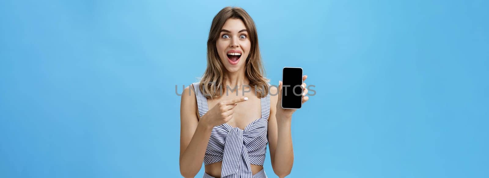 Excited happy woman with gapped teeth finally bought brand new smartphone holding device in hand pointing at cellphone screen showing cool app smiling broadly from joy against blue wall by Benzoix
