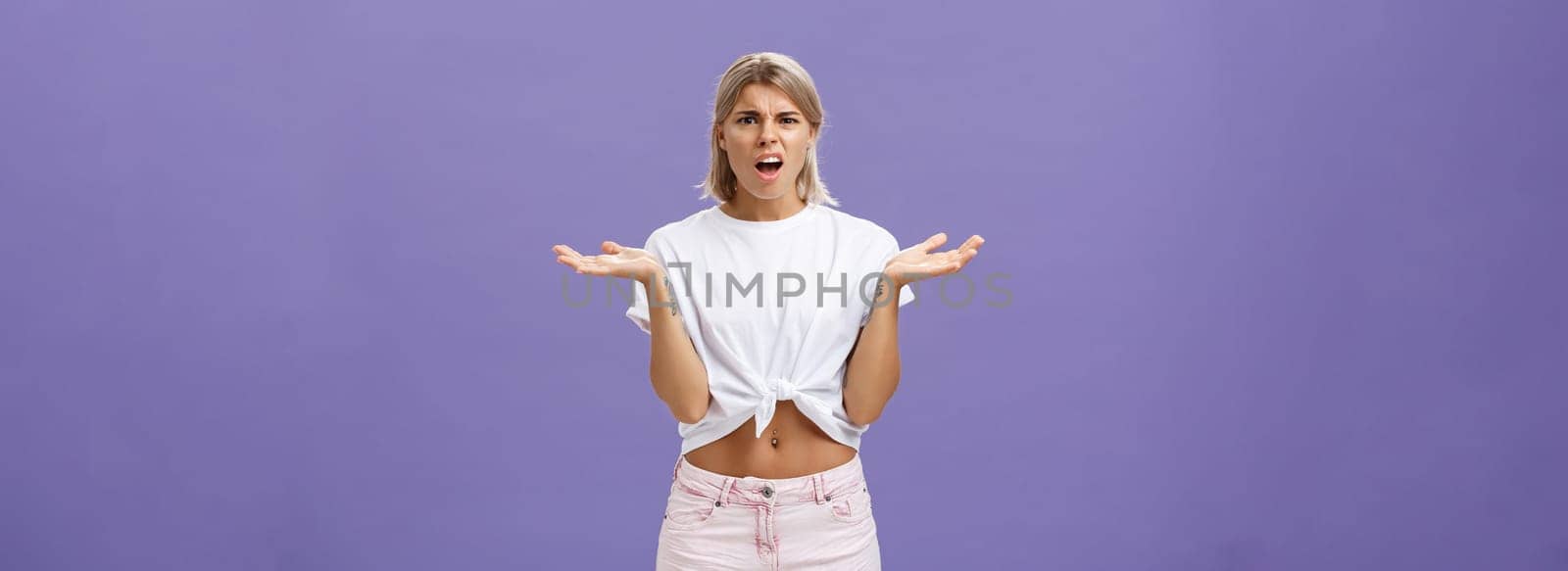 Perplexed and disappointment good-looking blonde female student in white t-shirt and pink shorts frowning shrugging with spread hands near shoulders saying what the hell over purple wall by Benzoix