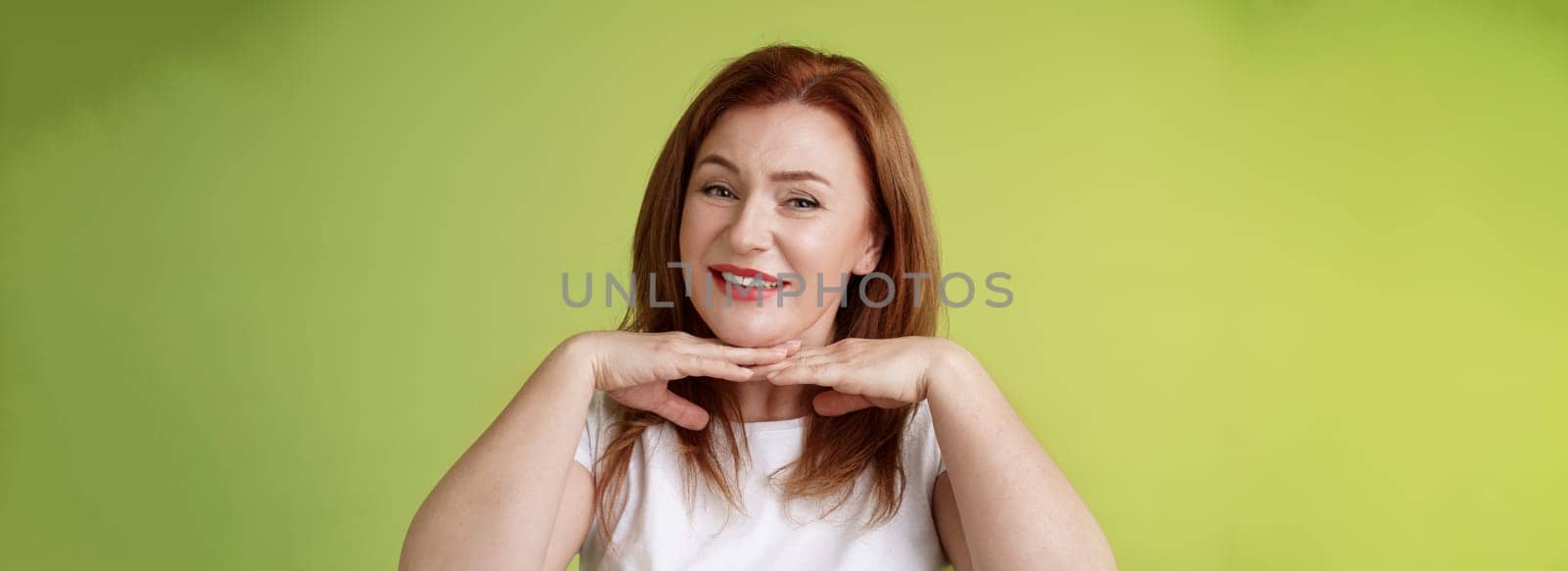 Looking good. Happy cheerful redhead middle-aged 50s woman smiling delighted hold hands under chin accept flaws blemished like own skin condition apply aging creme cosmetics green background.