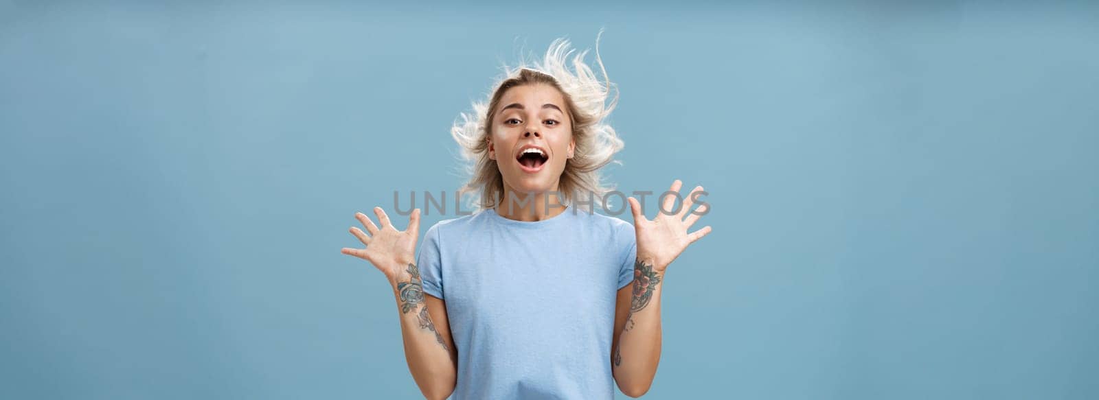 Creative happy and playful beautiful blond girl with tattoos on arms raising palms up opening mouth facing wind while hair strands flicking on air jumping having fun over blue background by Benzoix