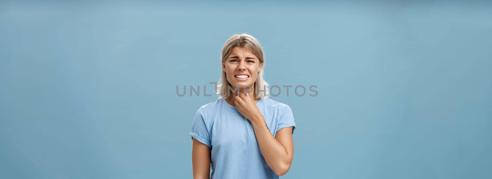 Girl feeling discomfort in throat catching cold or having seasonal allergy touching neck and frowning with clenched teeth and painful expression standing displeased over blue background. Health concept