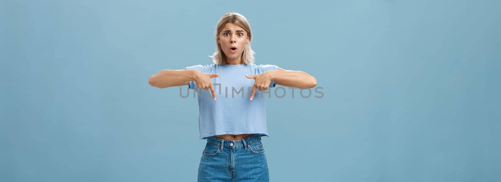 Studio shot of worried caucasian young woman feeling nervous her shoes do not fit frowning asking advice nervously pointing down shocked while frowning and standing over blue background by Benzoix