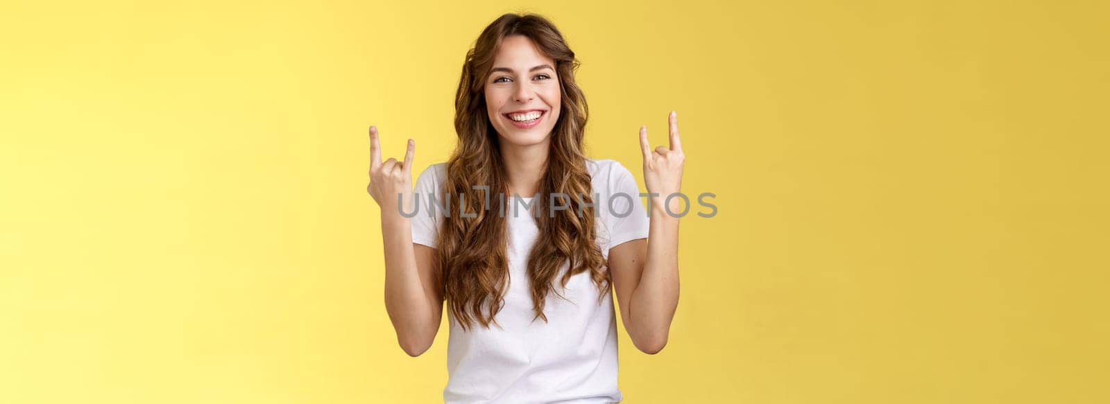 Happy cheerful feminine caucasian girl curly hairstyle show rock-n-roll heavy metal gesture smiling broadly enjoy awesome concert atmosphere stand yellow background joyfully cheering satisfied. Lifestyle.