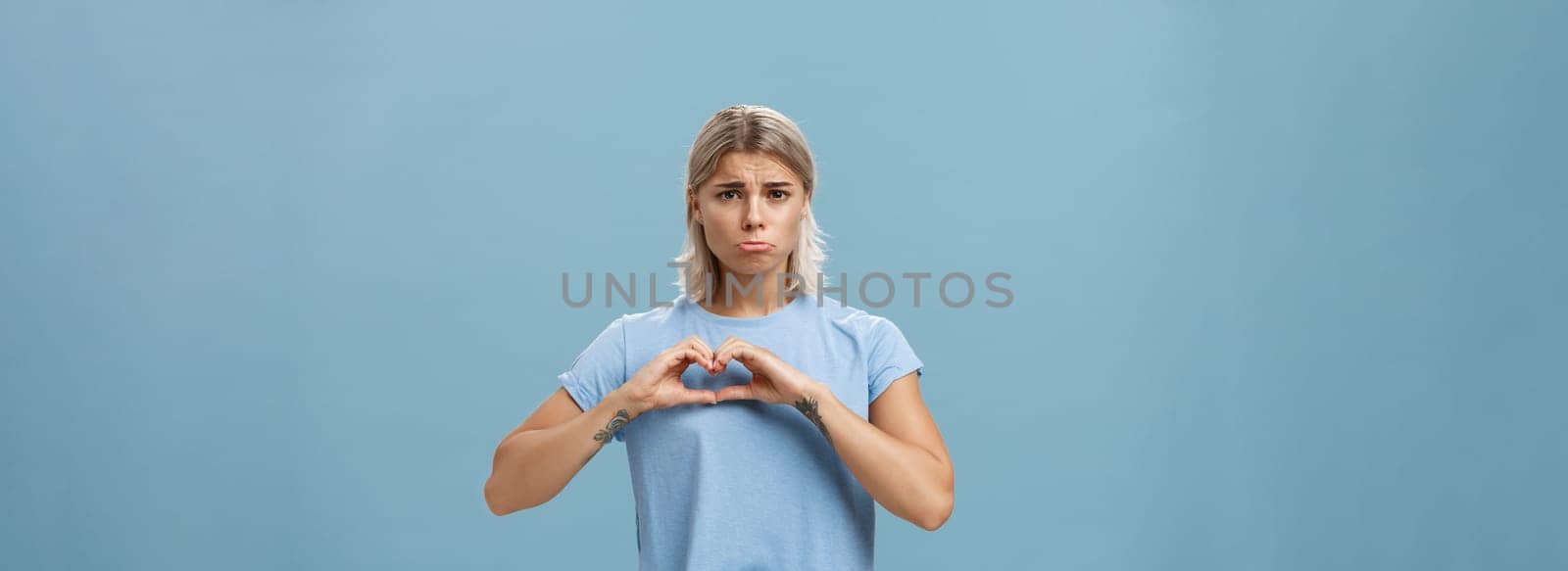 Heart being broken. Sad and gloomy heartbroken girl with blond hair tattoos on arms and tanned skin pursing lips whining and complaining making love sign over breast standing unhappy near blue wall by Benzoix