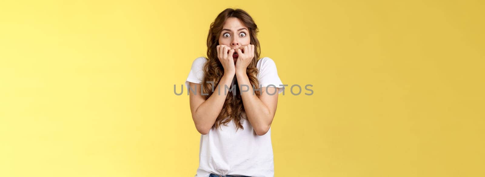 Shocked speechless scared stunned curly-haired caucasian woman gasping hold fingers mouth biting nails stare camera frightened express fear terrified scary movie stand yellow background.