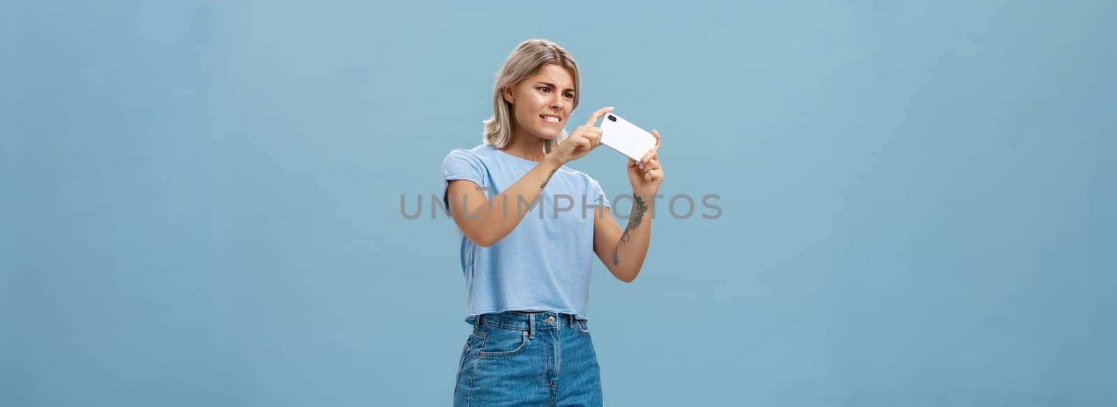 Girl trying to take photo of pet moving fast staring intense at smartphone screen pointing phone camera left feeling focused while taking picture on cellphone over blue background by Benzoix