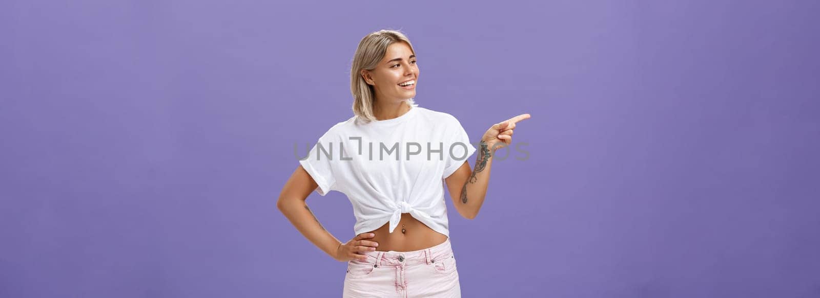 Hey nice job. Pleased carefree good-looking and confident female shop manager in stylish outfit holding hand on waist pointing and looking left with satisfied and delighted look over purple wall by Benzoix