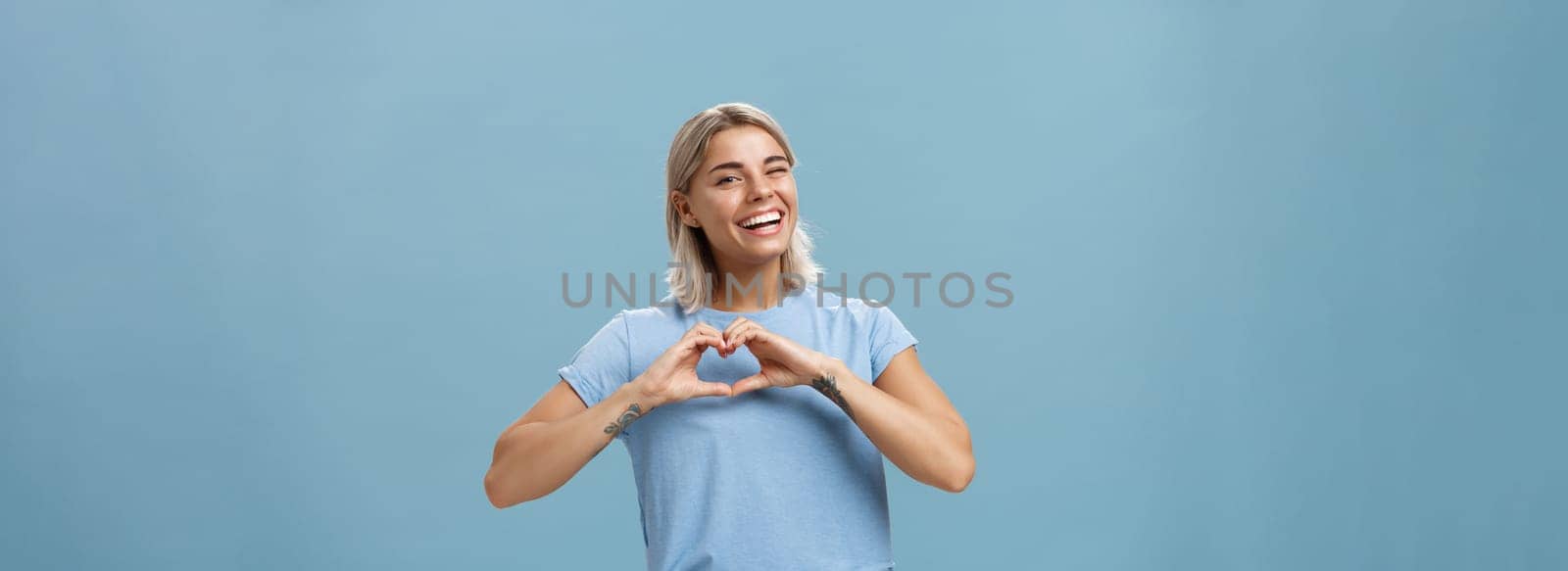 Love relationship and emotions concept. Joyful charming feminine girl with fair hair in trendy t-shirt winking happily smiling broadly and making heart gesture over breast expressing affection by Benzoix