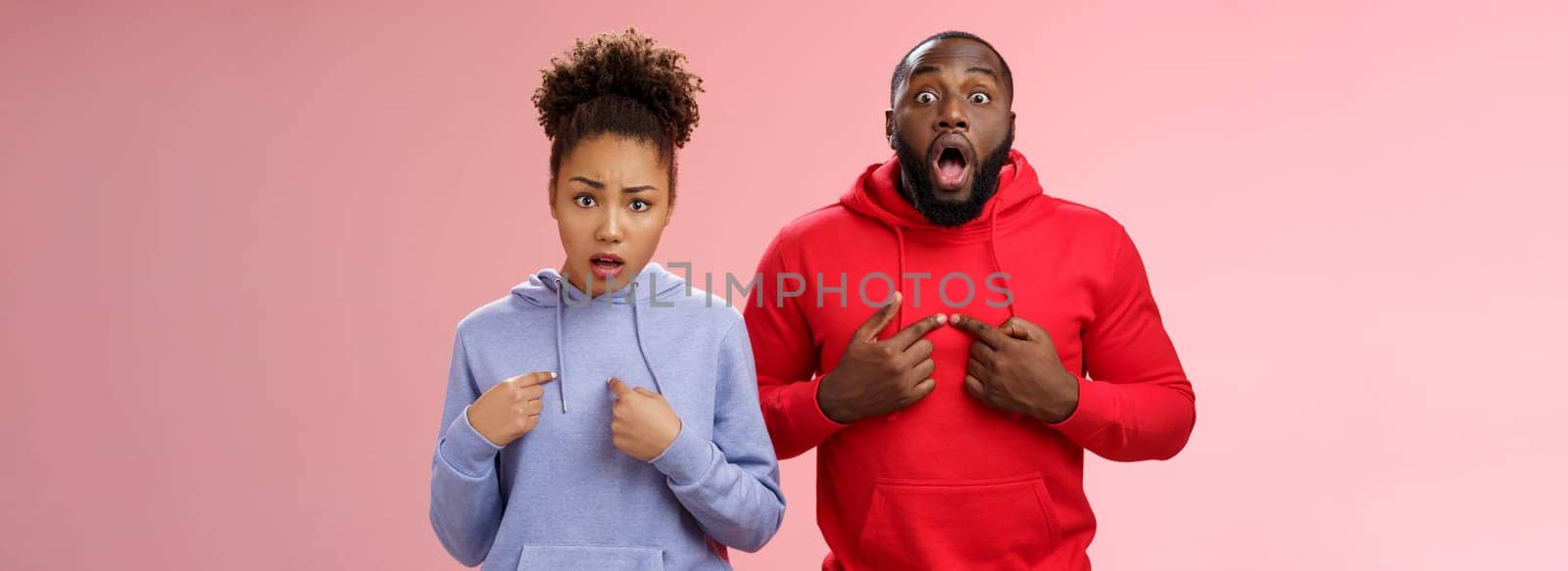 Why us. Shocked disturbed complaining two african american brother sister pointing chest index fingers frowning bothered upset man widen eyes surprised woman frowning sorrow, standing pink wall.