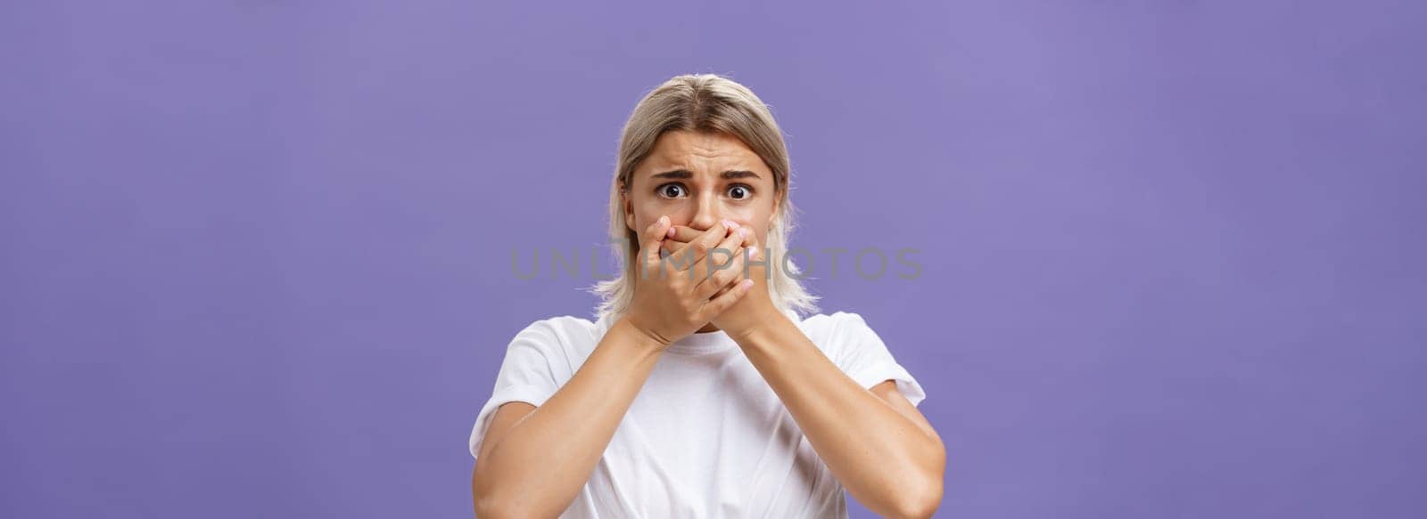 Waist-up shot of shocked nervous and scared young woman witnessing terrible crime covering mouth with both hands not to scream frowning staring frightened at camera over purple background. Emotions concept