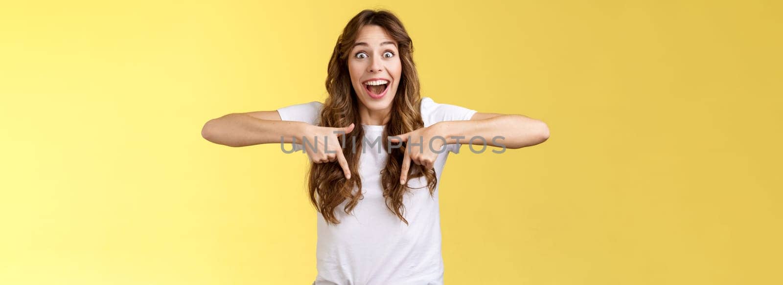 Excited happy cheerful impressed cute brunette girl long haircut smiling broadly surprised react pleased pointing down share excellent positive news stand joyfully yellow background promoting.