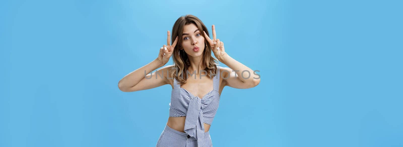 Charming cute unretouched woman with chestnut hair and tattoo showing peace gestures near face folding lips in mwah making silly face while posing over blue background confident and carefree. Emotions and body language concept