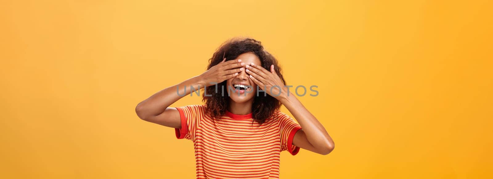 Closing my eyes and counting ten. Portrait of charming dreamy and happy funny african american curly-haired female in striped trendy t-shirt covering sight with palm and smiling waiting surprise by Benzoix