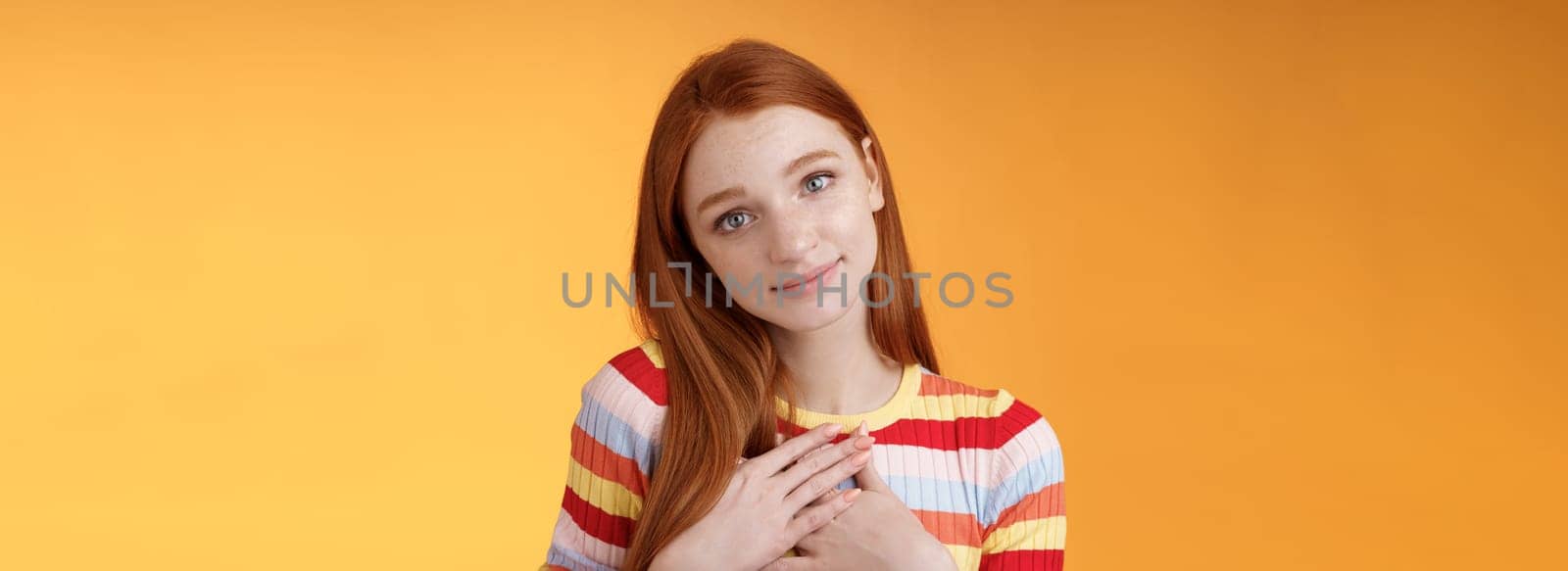 Lovely silly cute redhead young european girl tilting head look gently smiling tender camera press palms heart thankful receive nice gift gazing gratitude happiness, standing orange background by Benzoix