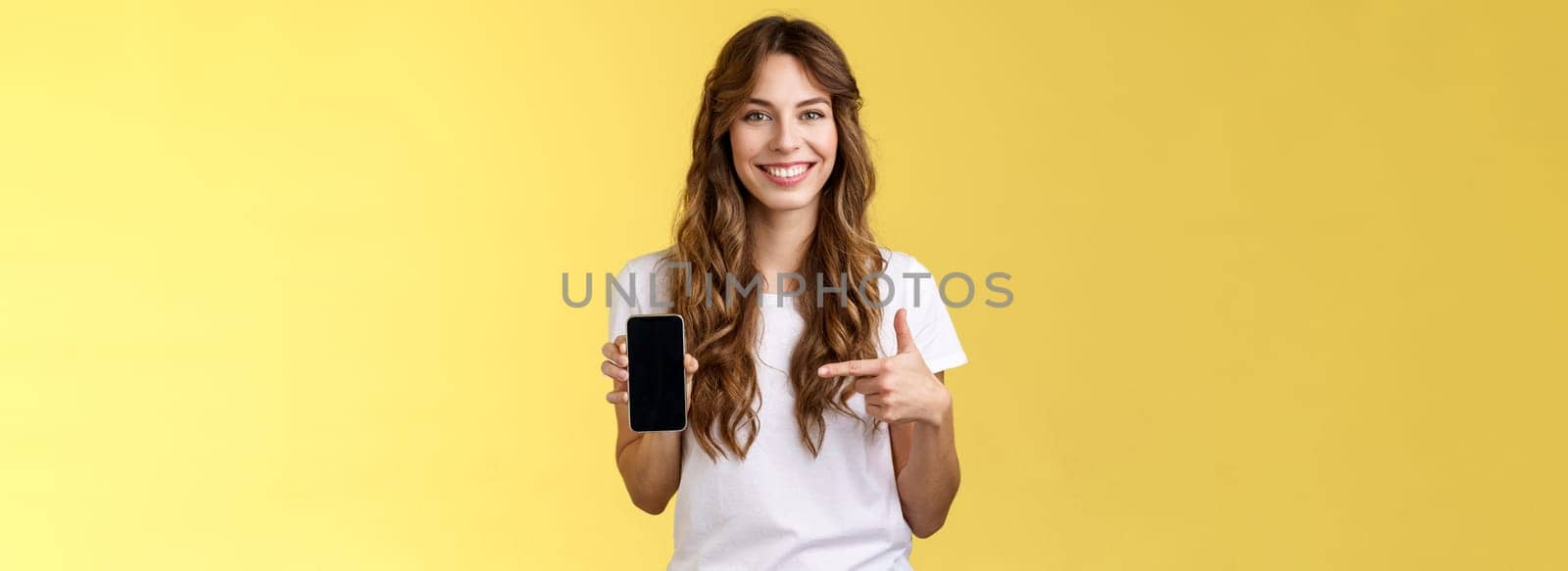 Girl have everything need smartphone. Cheerful relaxed carefree woman long curly hairstyle hold mobile phone pointing forefinger cellphone smiling broadly explain how app works yellow background.