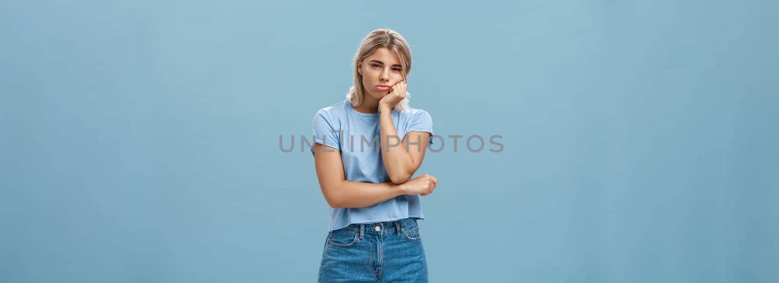 Studio shot of displeased moody girlfriend feeling disappointed boyfriend spoilt date leaning head on palm sulking from dissatisfaction and squinting being offended posing over blue background by Benzoix