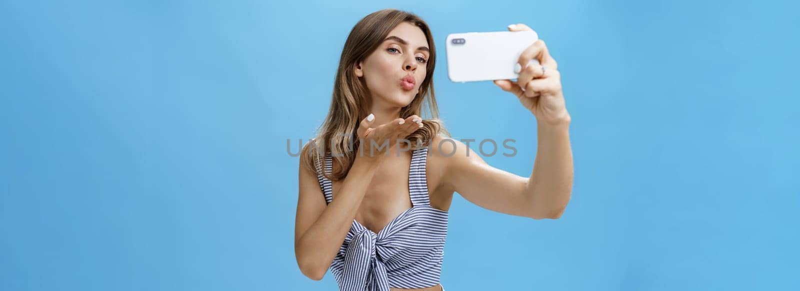 Outgoing and self-assured glamorous woman like taking pictures of herself holding smartphone making sensual and flirty selfie folding lips raising palm to send wind kiss at screen loving followers. Technology, lifestyle, social network concept