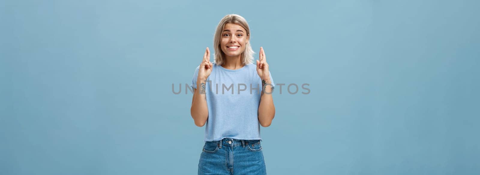 Hope dream come true soon. Portrait of emotive happy and optimistic attractive young european woman with blond hair and tanned skin crossing fingers for good luck and smiling over blue wall.