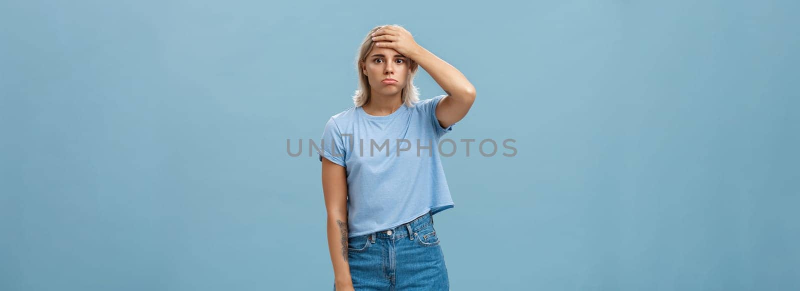 Concerned and troubled young teenage girl being shocked with loads of responsibilities holding hand on forehead staring spaced out and pouting from tiresome perplexed situation over blue wall by Benzoix