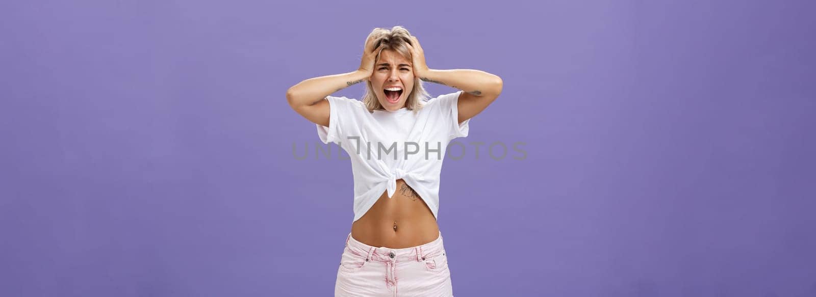 Portrait of panicking concerned and depressed young european woman with blond hair tattoos and athletic body yelling out loud holding hands on head being concerned and shook over purple background by Benzoix