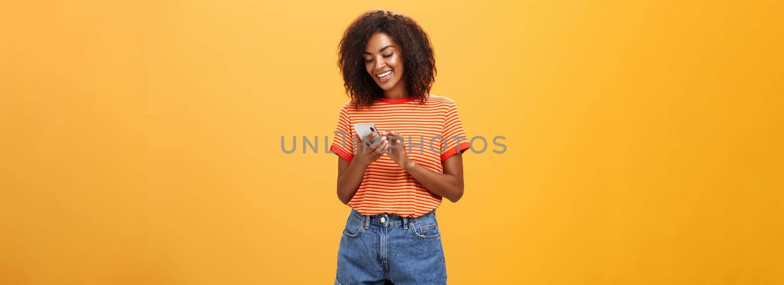 Stylish carefree girl texting friend come over standing pleased over orange wall in stylish denim shorts typing message or scrolling news in internet via smartphone gazing at device screen with smile. Lifestyle.